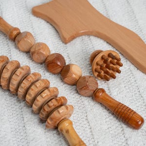 Pode incluir: Uma coleção de ferramentas de massagem de madeira, incluindo um rolo, um massageador de bolas, um pequeno massageador redondo com pinos e uma ferramenta de madeira curvada. As ferramentas são feitas de madeira marrom clara e estão dispostas em uma superfície branca.