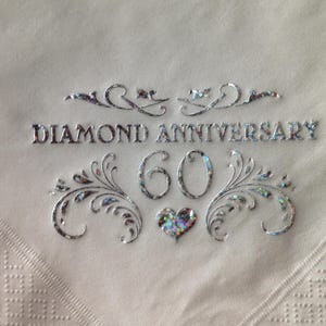 Puede incluir: Servilleta de color plateado con el texto "Diamond Anniversary 60" y un diseño de corazón decorativo.