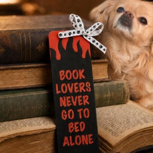 May include: A rectangular black bookmark with red flame-like patterns at the top and the red text "BOOK LOVERS NEVER GO TO BED ALONE" printed in the center. A white ribbon with small black paw prints is tied in a bow at the top of the bookmark.