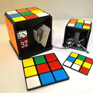 May include: A black and orange Rubik's Cube-shaped box with a hidden compartment. The box contains dice, a red die with the number 32, and a card. Additional Rubik's Cube-themed accessories are also present.