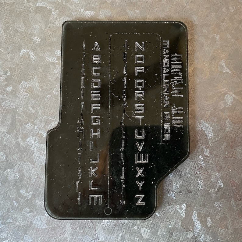 May include: Black plastic card with a Mandalorian alphabet guide. The card has a hole in the bottom right corner. The text on the card reads "Mandalorian Guide".