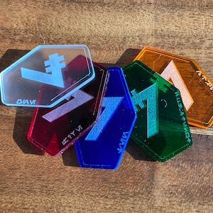 May include: Five hexagonal acrylic tokens in different colours and symbols. The tokens are red, blue, green, orange, and clear. The symbols are a stylised letter "V" with a vertical line, a stylised letter "V" with a horizontal line, a stylised letter "V" with a diagonal line, a stylised letter "V" with a curved line, and a stylised letter "V" with a straight line.