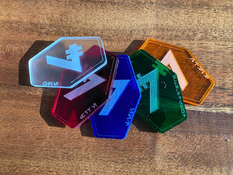 May include: Five hexagonal acrylic tokens in different colours and symbols. The tokens are red, blue, green, orange, and clear. The symbols are a stylised letter "V" with a vertical line, a stylised letter "V" with a horizontal line, a stylised letter "V" with a diagonal line, a stylised letter "V" with a curved line, and a stylised letter "V" with a straight line.