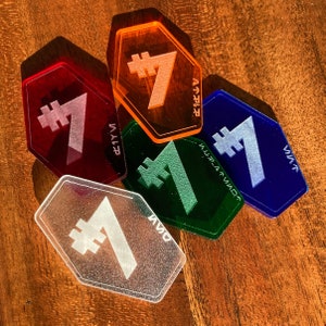 May include: Five hexagonal acrylic tokens, each with a different colour and a number seven with a hash symbol in front of it. The colours are orange, red, green, blue, and clear.