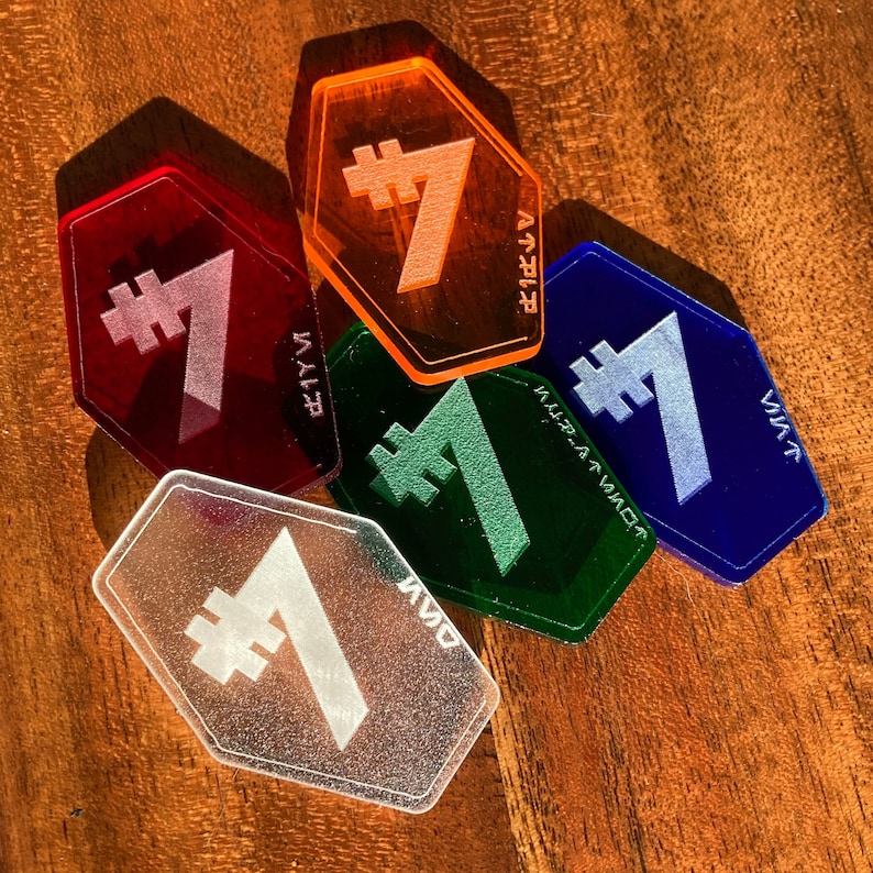 May include: Five hexagonal acrylic tokens, each with a different colour and a number seven with a hash symbol in front of it. The colours are orange, red, green, blue, and clear.