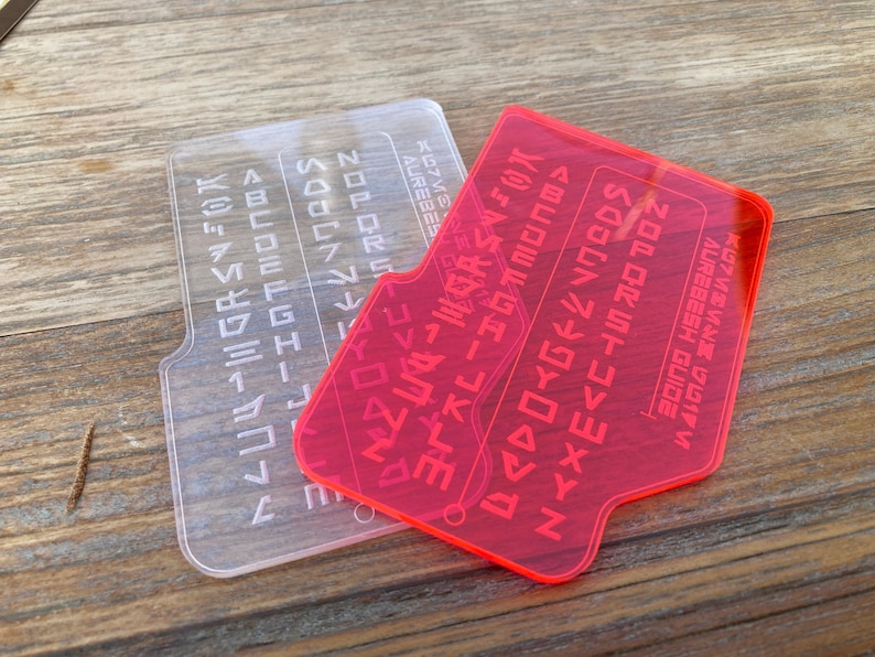 May include: Two plastic alphabet stencils, one clear and one pink, with a raised alphabet in a futuristic font. The stencils are on a wooden surface.