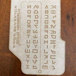 May include: A white card with a brown border and a brown circle in the center. The card has a grid of symbols, with the text "Aurebesh  Guide" and "Universal  Alphabet" on the left side.