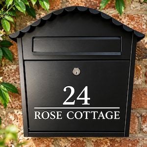May include: A matte black wall-mounted mailbox with a scalloped roof edge and a mail slot. The front panel features the white text 24 and ROSE COTTAGE centered above a horizontal line, with a central silver key lock mechanism for secure access.
