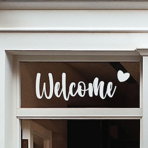Puede incluir: Ventana con la palabra "Welcome" en escritura blanca, acompañada de un corazón blanco. La ventana está enmarcada en blanco, sobre un fondo marrón oscuro.