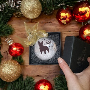 Op de afbeelding: Een transparante glazen kerstbal met een zwart silhouet van een Franse Bulldog en de naam "Milo" erop gedrukt. De bal zit in een zwarte cadeaudoos met een wit sneeuwvlokkenpatroon.