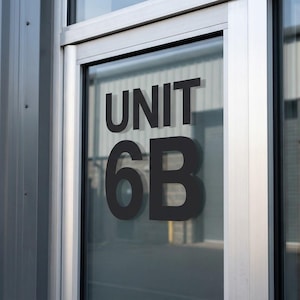May include: A glass door with the black text "UNIT 6B" displayed prominently. The door is set in a silver frame and reflects the surrounding industrial building. The overall color scheme is neutral, with shades of gray and silver.
