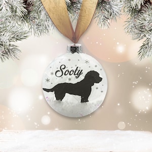 May include: White Christmas ornament with a black silhouette of a dog and the name "Sooty" written on it. The ornament is filled with faux snow.