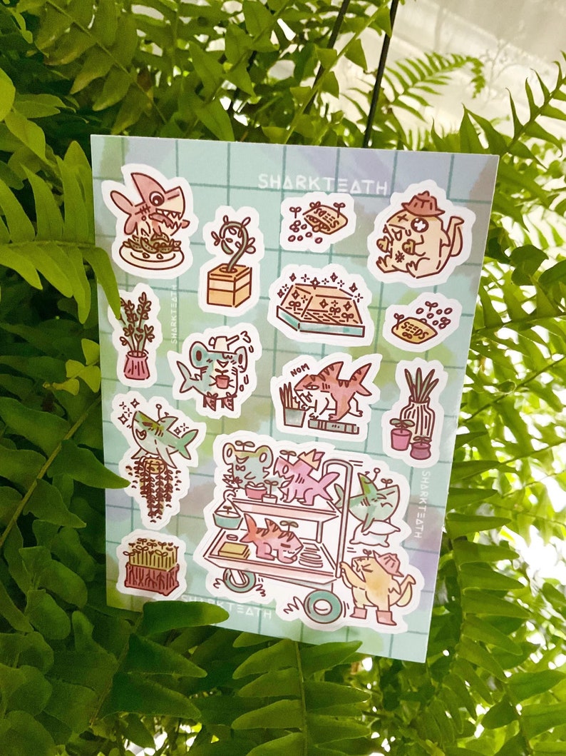 Puede incluir: Una hoja de pegatinas con lindas ilustraciones de dibujos animados de tiburones, plantas y otras criaturas marinas. Las pegatinas est&aacute;n sobre un fondo azul claro con un patr&oacute;n de cuadr&iacute;cula. El texto "SHARKTEATH" est&aacute; impreso en la esquina superior derecha de la hoja.
