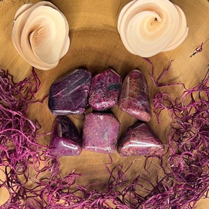 May include: Six tumbled purple ruby stones on a wooden plate with a pink and white background.
