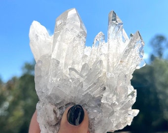 May include: A large cluster of clear quartz crystals with a rough, natural texture. The crystals are arranged in a pointed, spiky formation.