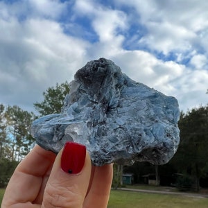 Puede incluir: Un cúmulo de cristales azul y gris con una textura rugosa y natural. El cristal se sostiene en una mano con esmalte de uñas rojo.