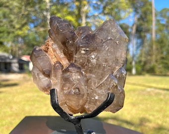 May include: A large, clear quartz crystal cluster with a smoky brown hue. The crystal is displayed on a black metal stand with a wooden base.