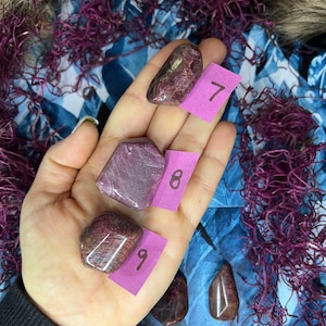 May include: Three pink and purple tumbled stones with white streaks, labeled 7, 8, and 9. The stones are on a hand with a purple background.