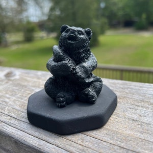 Könnte beinhalten: Eine schwarze Keramikfigur eines Bären, der mit gekreuzten Armen sitzt. Der Bär sitzt auf einem schwarzen sechseckigen Sockel.
