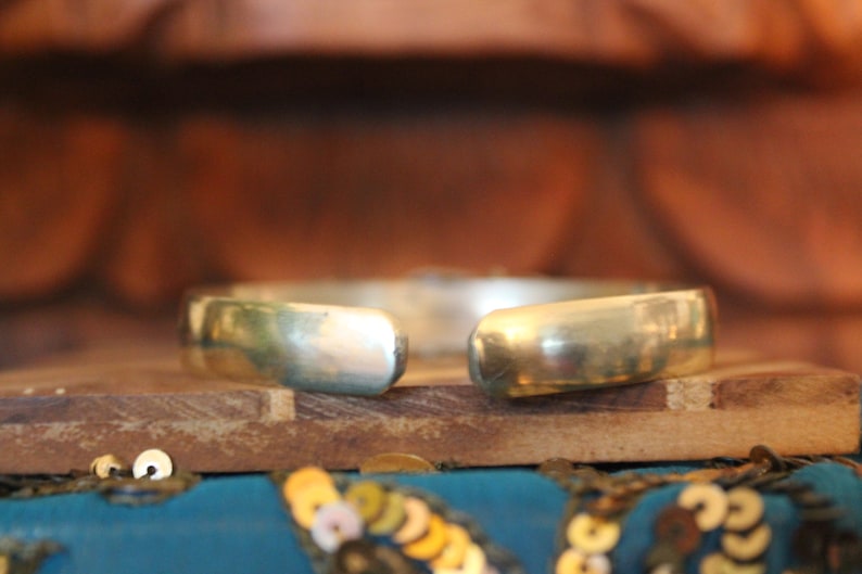 K&ouml;nnte beinhalten: Ein silbernes Armreif-Armband mit glatter, polierter Oberfl&auml;che. Das Armband ist an einer Seite offen und hat eine leicht gebogene Form.