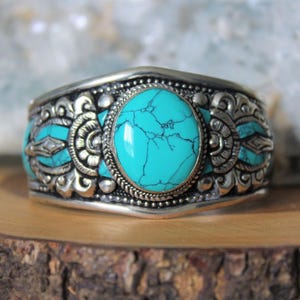 May include: Silver cuff bracelet with a large, oval turquoise stone at its centre. The bracelet features detailed, ornate patterns and smaller turquoise accents. The turquoise stone displays a distinctive, web-like pattern.
