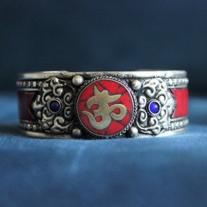 May include: Silver bracelet with red and blue accents. The bracelet features a circular design with the symbol "Om" in gold.