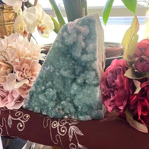 Puede incluir: Una geoda de cristal triangular grande, de color verde azulado. La geoda se exhibe sobre una superficie granate con bordados dorados, rodeada de flores rosas y blancas. Se ven cristales adicionales debajo.