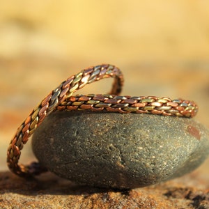 Puede incluir: Dos pulseras de cobre trenzadas con acabado dorado y plateado, descansando sobre una roca gris.