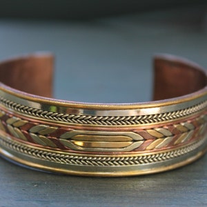 May include: A copper, silver, and gold bracelet with a braided design. The bracelet is wide and has a rounded shape.