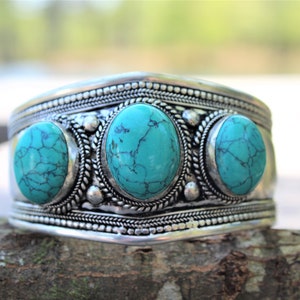 May include: A silver cuff bracelet with three turquoise stones set in the center. The bracelet has a textured design and is decorated with small beads.