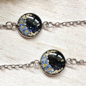 Puede incluir: Dos pulseras de cadena plateada con dijes redondos de vidrio. Cada dije presenta un fondo negro con flores azules y blancas y purpurina plateada.