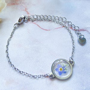 Puede incluir: Pulsera plateada con un dije redondo que contiene flores secas azules y blancas encapsuladas en resina transparente. La pulsera tiene una cadena delicada y un pequeño dije en forma de corazón en el cierre.