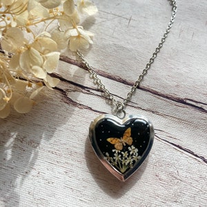 Puede incluir: Un collar con colgante en forma de corazón plateado. El colgante presenta un fondo negro con una mariposa dorada y pequeñas flores blancas. El collar está sobre una cadena plateada. El fondo es una superficie de madera marrón claro.