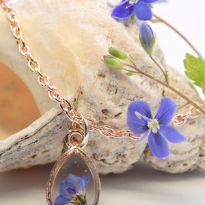 Puede incluir: Un delicado collar de oro con un colgante en forma de l&aacute;grima que presenta una flor azul prensada. El colgante est&aacute; rodeado por un marco de oro y cuelga de una delicada cadena de oro.