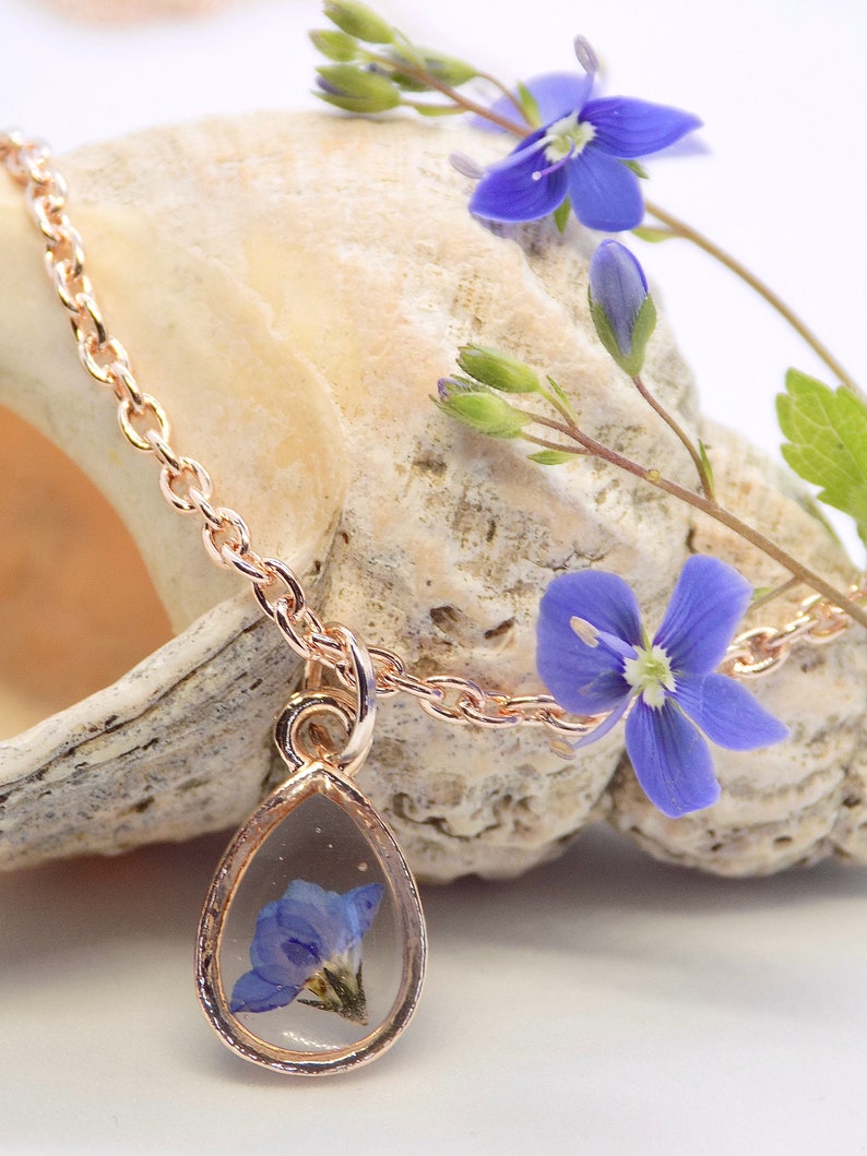 Puede incluir: Un delicado collar de oro con un colgante en forma de l&aacute;grima que presenta una flor azul prensada. El colgante est&aacute; rodeado por un marco de oro y cuelga de una delicada cadena de oro.