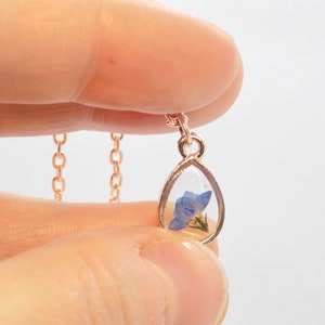 Puede incluir: Un delicado collar de cadena dorada con un colgante en forma de l&aacute;grima. El colgante est&aacute; relleno de resina transparente y contiene una flor azul prensada.