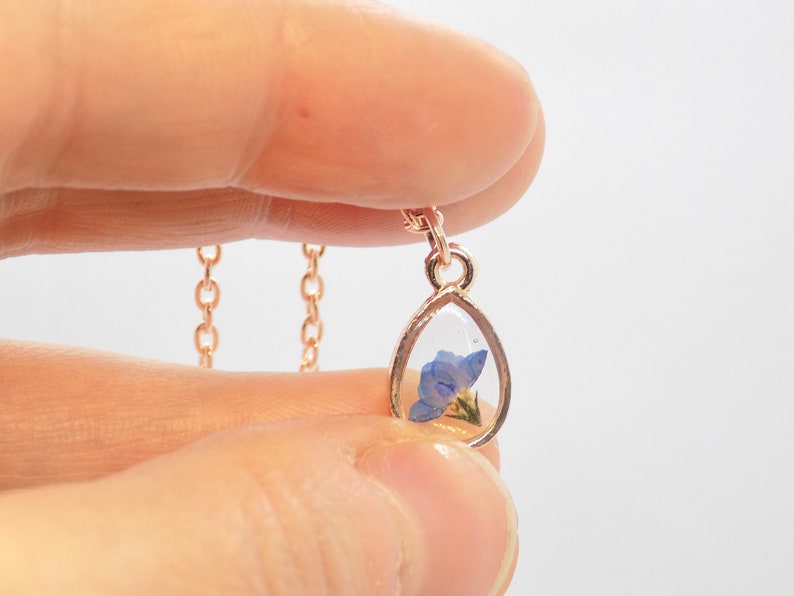 Puede incluir: Un delicado collar de cadena dorada con un colgante en forma de l&aacute;grima. El colgante est&aacute; relleno de resina transparente y contiene una flor azul prensada.