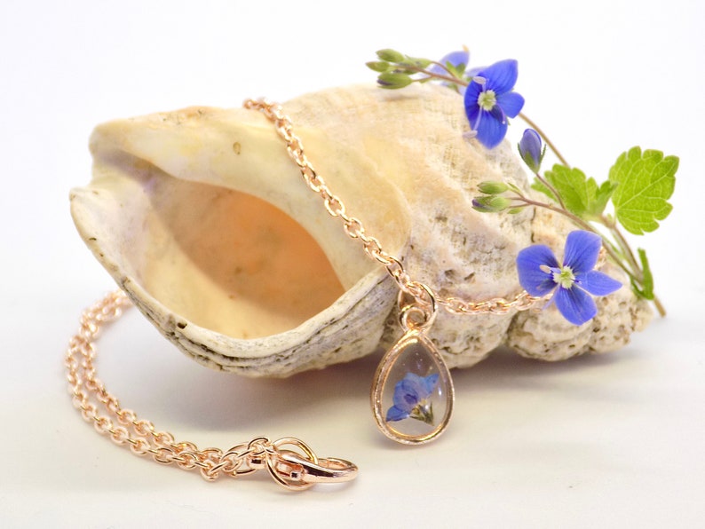 Puede incluir: Un delicado collar de oro con un colgante en forma de l&aacute;grima que presenta una flor azul prensada. El collar se muestra en una concha de mar con una ramita de flores azules.