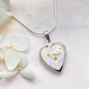 May include: A silver necklace with a heart-shaped pendant. The pendant contains small, light blue and white flowers with yellow centres, and a small pearl. The pendant is approximately 2.5 cm wide.