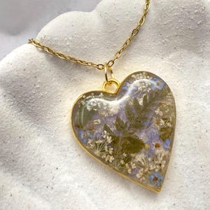 Könnte beinhalten: Halskette mit herzförmigem Anhänger und goldfarbener Kette. Der Anhänger ist mit getrockneten Blumen in Grün, Weiß und Blau gefüllt, eingebettet in klarem Harz. Der Anhänger hat einen goldenen Rand.