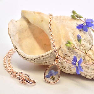 Puede incluir: Un delicado collar de oro rosa con un colgante en forma de l&aacute;grima que contiene una flor azul prensada incrustada en resina transparente.