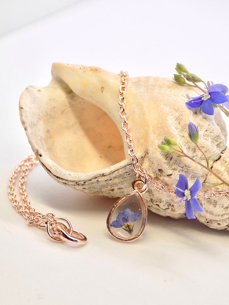 Puede incluir: Un delicado collar de oro rosa con un colgante en forma de l&aacute;grima que contiene una flor azul prensada incrustada en resina transparente.