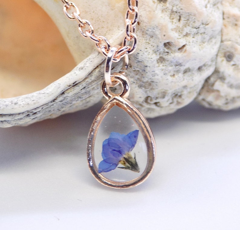 Puede incluir: Un delicado collar de oro con un colgante en forma de l&aacute;grima que presenta una flor azul prensada encapsulada en resina transparente.