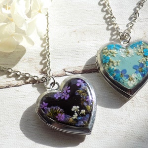 Puede incluir: Dos colgantes de plata en forma de corazón con cadenas. Un colgante tiene un fondo azul con flores blancas y azules. El otro colgante tiene un fondo negro con flores moradas y blancas.
