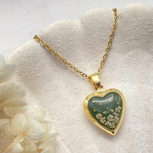 以下が含まれることがあります： 金色のハート型ペンダントネックレス。ペンダントは、小さな白い花と金のアクセントが付いた緑色の樹脂の中心部を持っています。ネックレスは金のチェーンで、背景はテクスチャ加工された白い表面です。