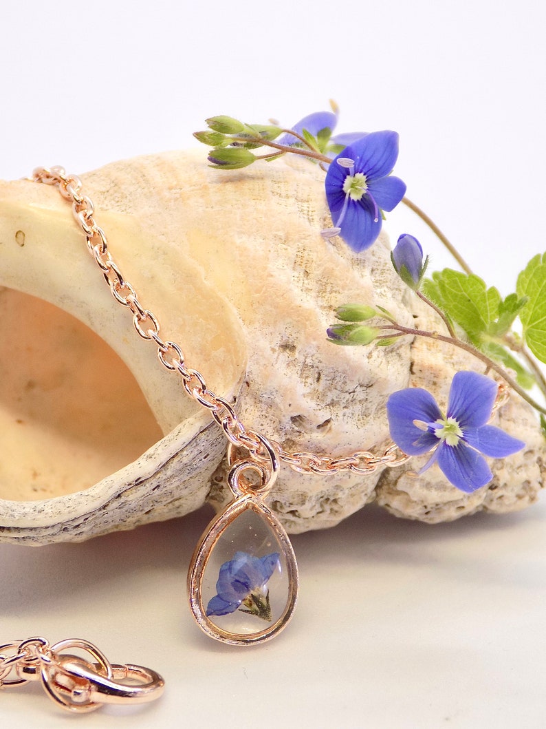 Puede incluir: Un delicado collar de oro rosa con un colgante en forma de l&aacute;grima que presenta una flor azul prensada. El collar se muestra en una concha de mar con una flor azul.