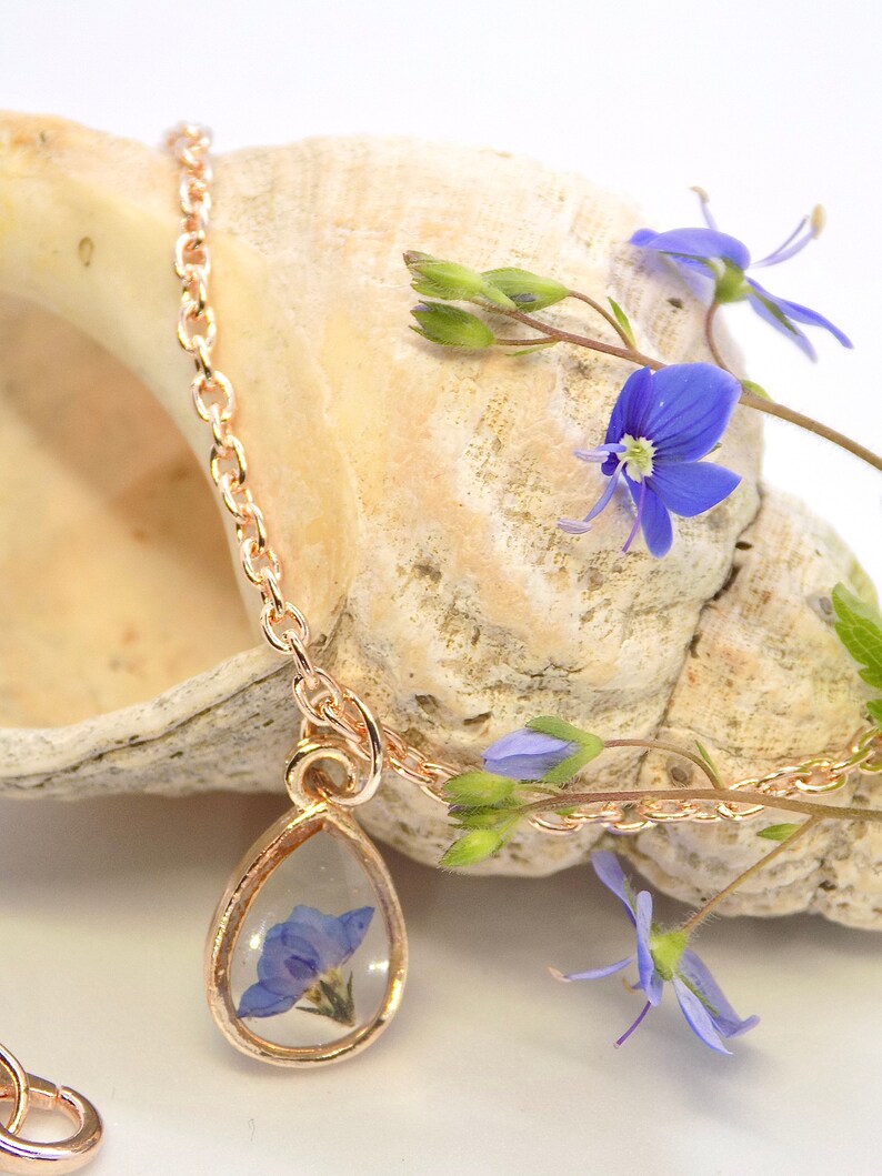 Puede incluir: Un delicado collar de cadena dorada con un colgante en forma de l&aacute;grima que presenta una flor azul prensada encapsulada en resina transparente. El collar se muestra en una concha de mar con flores azules.