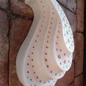 May include: White ceramic wall sculpture with a swirling, organic shape and a pattern of small holes. The sculpture is mounted on a brick wall.