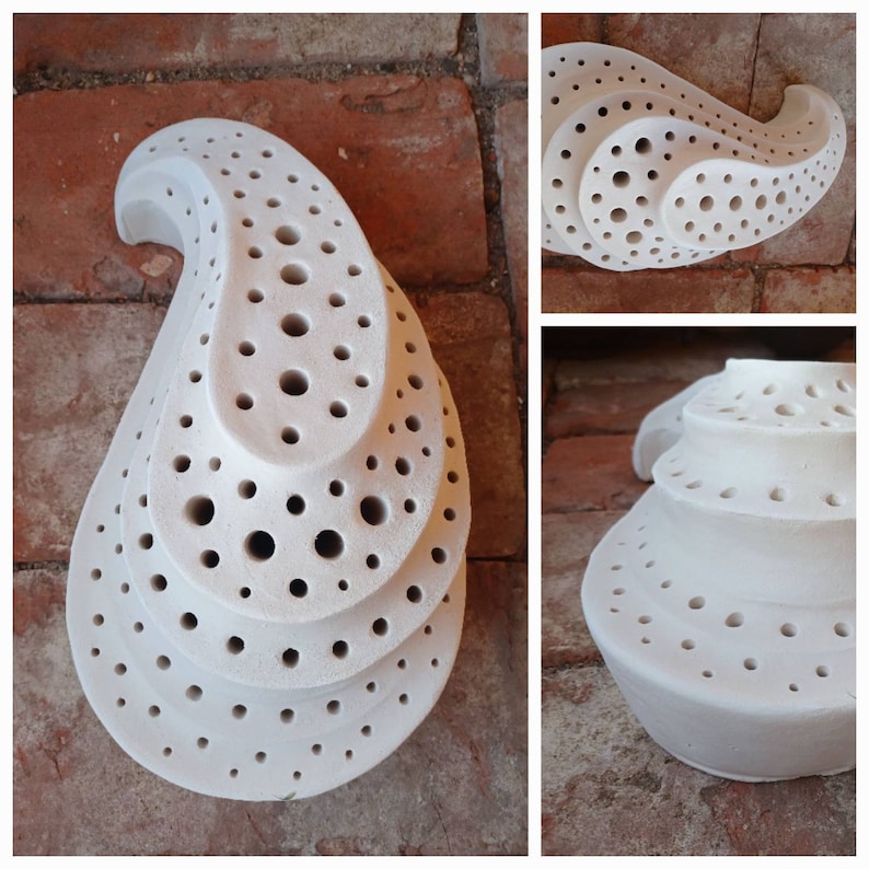 May include: White ceramic sculpture with a teardrop shape and multiple layers of holes. The sculpture is made of a smooth, white ceramic and has a unique, organic design.