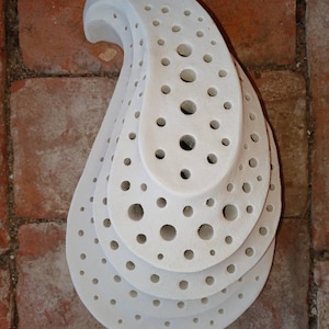 May include: White ceramic teardrop-shaped object with multiple layers of circular holes. The object is made of white ceramic and has a smooth, matte finish.